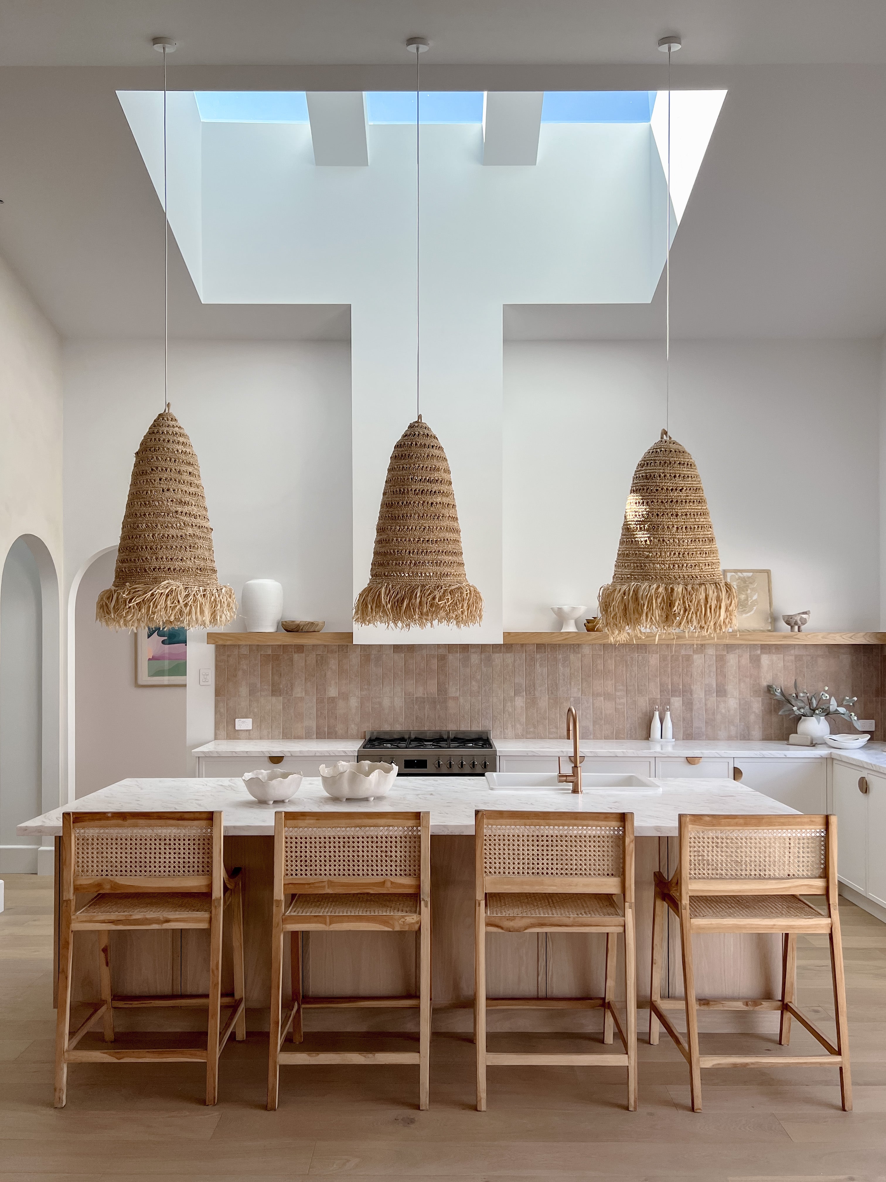 A coastal and open plan kitchen with a huge skylight and three rattan pendant lights are hanging over the island bench